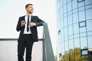 barbudo homem de negocios dentro formal terno em pausa usando Móvel telefone usar Smartphone. o negócio homem em pé lado de fora em moderno urbano cidade rua fundo com café copo dentro centro da cidade ao ar livre. cópia de espaço foto