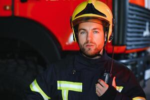 confiante bombeiro vestindo protetora uniforme em pé Próximo para uma fogo motor dentro uma garagem do uma fogo departamento foto