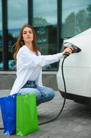 feliz jovem mulher em pé em cidade estacionamento perto elétrico carro, cobrando automóvel bateria a partir de pequeno cidade estação, segurando compras bolsas. foto