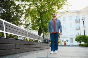 jovem cego homem com branco bengala caminhando através a rua dentro cidade foto