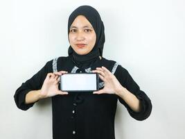 jovem muçulmano mulher dentro hijab sorridente mostrando Móvel telefone em branco tela recomendando aplicativo foto