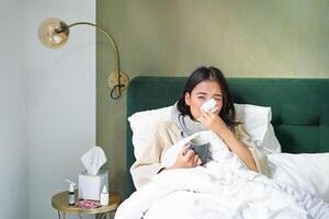 COVID-19, gripe e vacinação conceito. coreano menina deitado dentro cama com frio, pegando gripe, bebendo quente chá e usando prescrito medicação foto