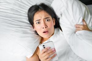 retrato do ásia menina acorda acima dentro manhã, parece às Móvel telefone com chocado face, dormiu demais, abraçando travesseiro e fazendo careta foto