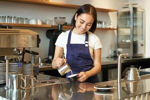 fofa coreano barista garota, derramando leite dentro café, preparar cappuccino com café com leite arte, trabalhando dentro cafeteria atrás contador. pessoas e local de trabalho foto