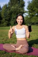 vertical tiro do ásia mulher mostra polegares acima, recomendando ioga Treinamento on-line, meditação aplicativo, fazendo exercícios em fresco ar dentro parque, mostrando Smartphone tela foto