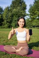 vertical tiro do ásia mulher mostra polegares acima, recomendando ioga Treinamento on-line, meditação aplicativo, fazendo exercícios em fresco ar dentro parque, mostrando Smartphone tela foto