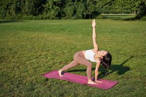 retrato do jovem ásia mulher alongamento, fazendo ioga em borracha esteira, exercício dentro parque, atento Treinamento em fresco ar foto