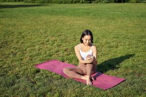 mulher dentro parque, assistindo ioga vídeo em Smartphone, meditando em fresco ar, sentado em borracha esteira foto