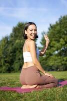 retrato do ásia menina em ginástica classe dentro parque, sentado em borracha esteira e onda mão às Câmera, dizer olá, meditando e prática ioga foto