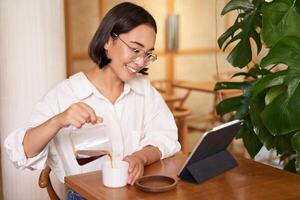 freelance e controlo remoto trabalhadores. sorridente jovem mulher derramando café dentro uma xícara, sentado dentro cafeteria e olhando às digital tábua foto