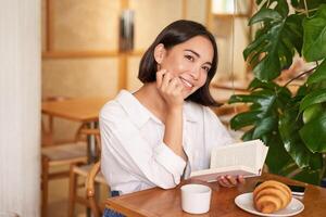 romântico ásia mulher sentado com livro dentro cafeteria, comendo croissant e bebendo café, lendo e sorridente, desfrutando sozinho Tempo foto