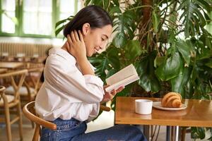 macio, lindo ásia menina sentado com uma livro dentro cafeteria, lendo e bebendo café. pessoas e estilo de vida conceito foto