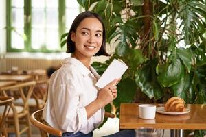 macio, lindo ásia menina sentado com uma livro dentro cafeteria, lendo e bebendo café. pessoas e estilo de vida conceito foto