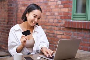 sorridente ásia mulher sentado com computador portátil, pagando de crédito cartão para conectados compras, enviando dela banco conta detalhes, sentado dentro cafeteria foto