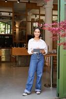 retrato do ásia mulher, Gerente em pé com tábua dentro frente do cafeteria Entrada, congratula-se com convidados foto