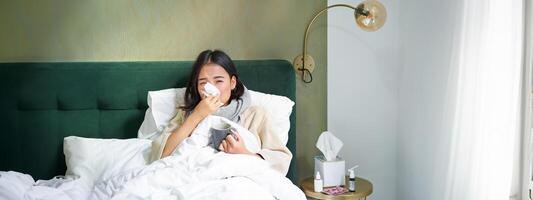 COVID-19, gripe e vacinação conceito. coreano menina deitado dentro cama com frio, pegando gripe, bebendo quente chá e usando prescrito medicação foto