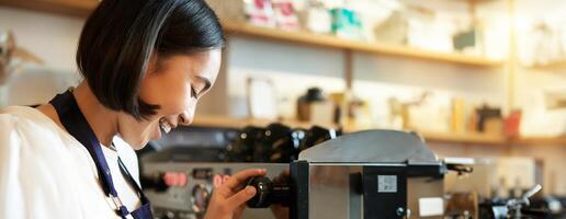 feliz ásia mulher, barista usando café máquina para faço ordem, vapor leite para cappuccino e café com leite, rindo e sorridente enquanto trabalhando dentro cafeteria foto