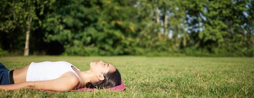 jovem ginástica menina deitado em esporte esteira em grama, respiração e meditando dentro parque dentro roupa de esporte foto