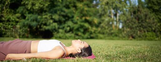 jovem ginástica menina deitado em esporte esteira em grama, respiração e meditando dentro parque dentro roupa de esporte foto