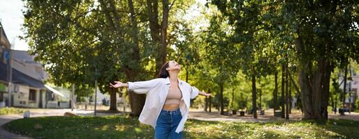 despreocupado ásia menina dançando, sentindo-me felicidade e alegria, desfrutando a Sol em verão dia, caminhando dentro parque com verde árvores foto