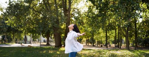 despreocupado ásia menina dançando, sentindo-me felicidade e alegria, desfrutando a Sol em verão dia, caminhando dentro parque com verde árvores foto