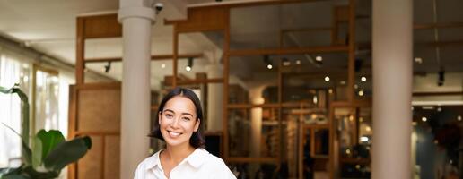 sorridente ásia gerente, confiante mulher em pé perto restaurante Entrada, cafeteria proprietário congratula-se com convidados foto