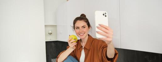 retrato do feliz, sorridente jovem mulher registros ela mesma, leva selfie enquanto comendo a maçã dentro a cozinha, usando Smartphone aplicativo, faz fotos com Móvel telefone