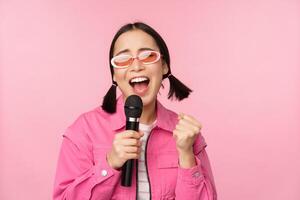 feliz lindo ásia menina cantando com microfone, usando microfone, desfrutando karaokê, posando contra Rosa estúdio fundo foto