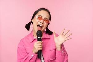 feliz lindo ásia menina cantando com microfone, usando microfone, desfrutando karaokê, posando contra Rosa estúdio fundo foto