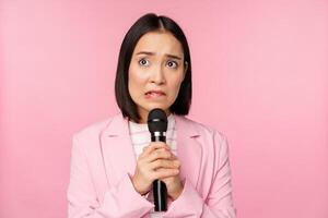 ansioso ásia senhora dentro terno, falando dentro público, dando discurso com microfone em conferência, olhando assustado, em pé sobre Rosa fundo foto