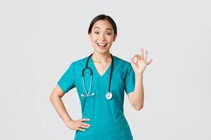 COVID-19, cuidados de saúde trabalhadores, pandemia conceito. surpreso e feliz ásia fêmea doutor, enfermeira dentro esfrega mostrando OK gesto e sorridente espantado, elogio agradável trabalhar, aceita com alguém foto