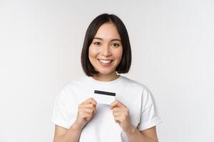 sorridente coreano mulher mostrando crédito cartão com feliz face, em pé dentro camiseta sobre branco fundo foto