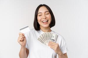 retrato do ásia mulher sorridente, segurando crédito cartão e dinheiro dinheiro, dólares, em pé dentro camiseta sobre branco fundo foto
