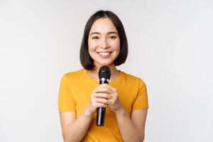 imagem do jovem ásia mulher falando dentro microfone, executar com microfone, dando discurso, em pé dentro amarelo camiseta contra branco fundo foto