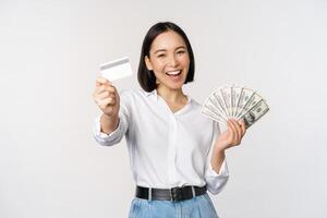 feliz coreano mulher segurando crédito cartão e dinheiro dólares, sorridente e rindo, posando contra branco estúdio fundo foto