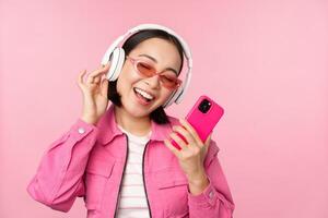 à moda ásia menina dançando com Smartphone, ouvindo música dentro fones de ouvido em Móvel telefone aplicativo, sorridente e rindo, posando contra Rosa fundo foto