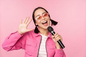 feliz lindo ásia menina cantando com microfone, usando microfone, desfrutando karaokê, posando contra Rosa estúdio fundo foto