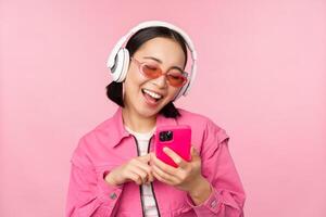 à moda ásia menina dançando com Smartphone, ouvindo música dentro fones de ouvido em Móvel telefone aplicativo, sorridente e rindo, posando contra Rosa fundo foto