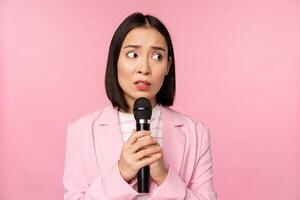 inseguro ásia empresária dando discurso, assustado do falando dentro público, usando microfone, em pé dentro terno sobre Rosa fundo foto