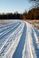 inverno panorama. uma enrolamento deserta estrada corrida frente através uma campo. coberto de neve floresta dentro a pôr do sol raios do a inverno Sol. carro faixas dentro a neve foto