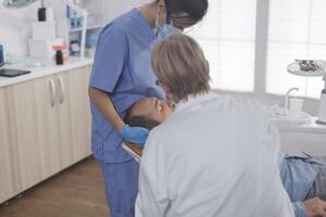 clínico ortodontista equipe com face mascarar examinando paciente boca durante médico cirurgia dentro estomatológico escritório sala. Senior mulher médico usando profissional odontologia Ferramentas para cavidade procedimento foto