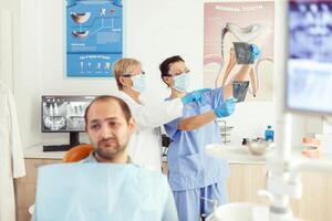 doente homem paciente esperando para dentes exame sentado em dental cadeira dentro estomatologia clínica sala. estomatologista médico e médico enfermeira preparando para ortodôntico cirurgia enquanto examinando radiografia foto