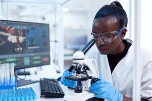 médico investigador com africano etnia usando microscópio fazendo investigação e avalia amostra. Preto cuidados de saúde cientista dentro bioquímica laboratório vestindo estéril equipamento. foto