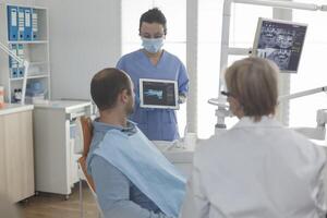 dentista assistente vestindo face mascarar mostrando dentes radiografia usando tábua para paciente discutindo médico procedimento durante estomatologia exame dentro dental escritório. doente homem tendo oral infecção foto