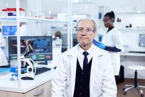 Senior masculino químico olhando às Câmera vestindo protetora óculos. idosos cientista vestindo laboratório casaco trabalhando para desenvolve uma Novo médico vacinar com africano assistente dentro a fundo. foto