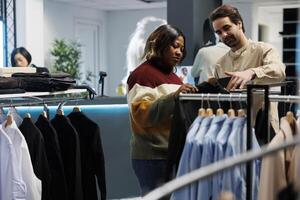 roupas loja cliente navegando prateleira com vestuário e falando com assistente. africano americano mulher obtendo Socorro a partir de boutique empregado dentro escolhendo na moda roupas enquanto compras foto