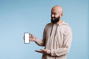 sorridente árabe anunciante apresentando Novo Smartphone com em branco branco tela zombar acima. alegre jovem pessoa segurando Móvel telefone dentro vertical modo com esvaziar tela sensível ao toque para promoção foto