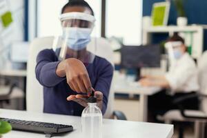 fechar acima do africano mulher usando mão desinfetante durante global pandemia com coronavírus. empresária dentro Novo normal local de trabalho desinfecção enquanto colegas trabalhando dentro fundo. foto