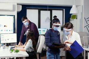 africano e caucasiano colegas de trabalho discutindo Estatisticas vestindo face mascarar contra covid19 às ambiente de trabalho. multiétnico o negócio equipe trabalhando respeitando social distância durante global pandemia com coronavírus. foto