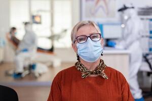 retrato do aposentado paciente dentro dental escritório olhando em Câmera vestindo face mascarar sentado em cadeira dentro esperando quarto clínica enquanto médico trabalhando. conceito do Novo normal dentista Visita dentro coronavírus surto. foto